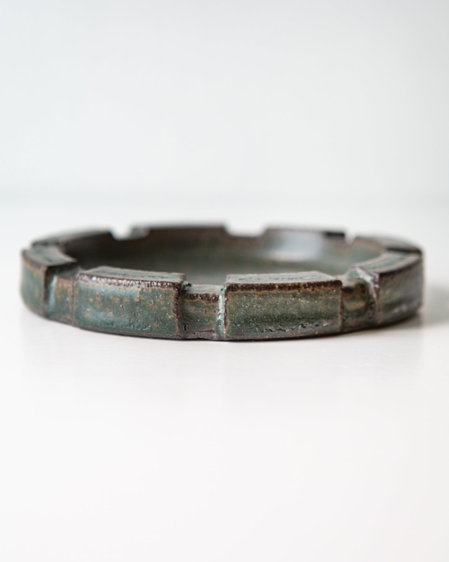 A close up shot of the side of a round ceramic ashtray with a brutalist carved rim and a speckled dark green glaze, shown on a clean white background.