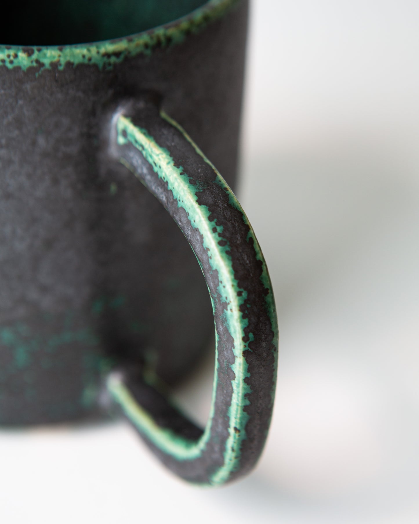 A close up shot of the handle of the ceramic mug with a textured matte black exterior, and a dark green glaze coloring on the edges, shown on a white background.