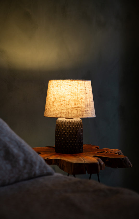 A dark brown hand carved table lamp with an off-white lamp shade, sitting on a wooden side table against a green wall. The light Is on showing a warm glow on the lamp shade.