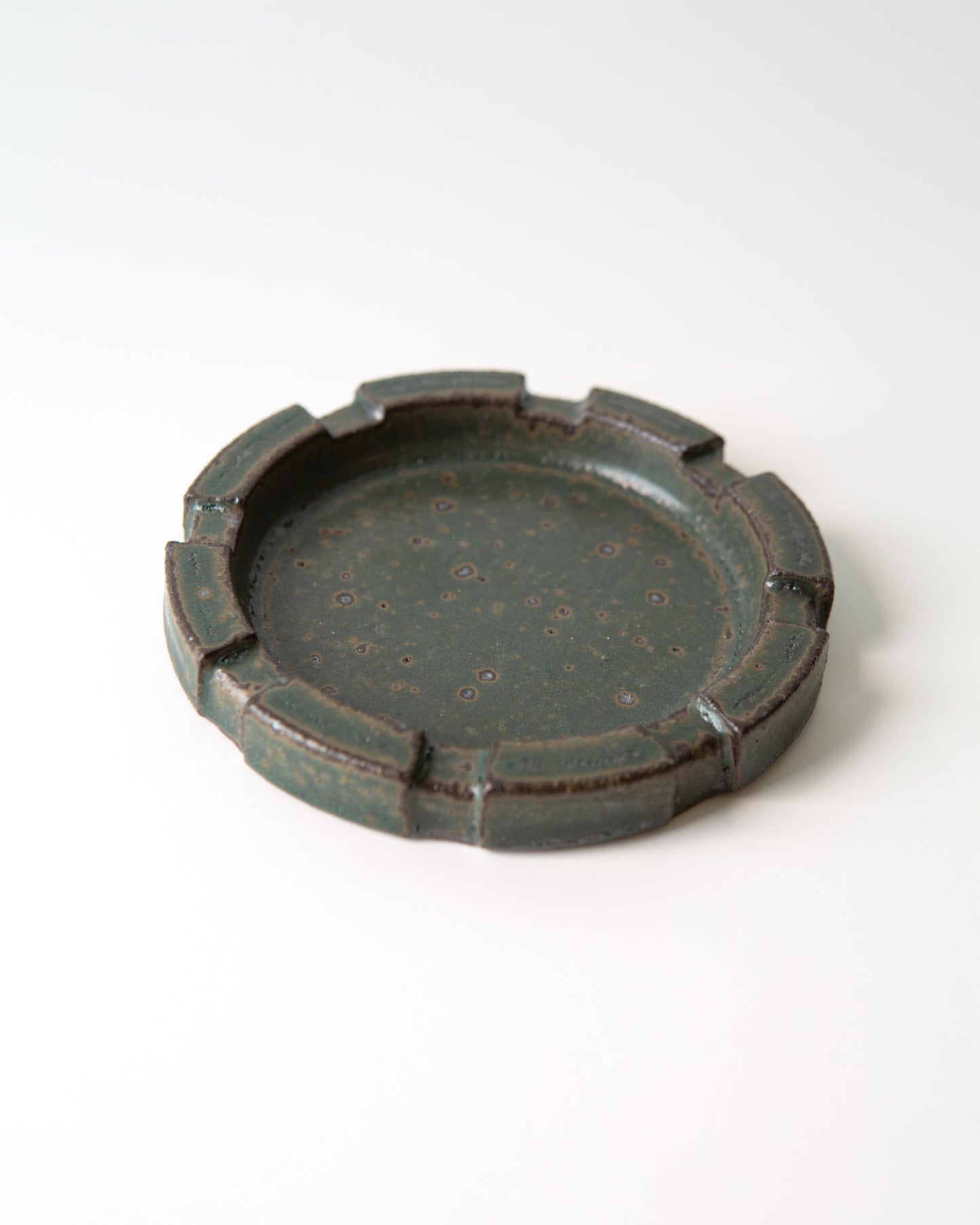 A round ceramic ashtray with a brutalist carved rim and a speckled dark green glaze, shown on a clean white background.