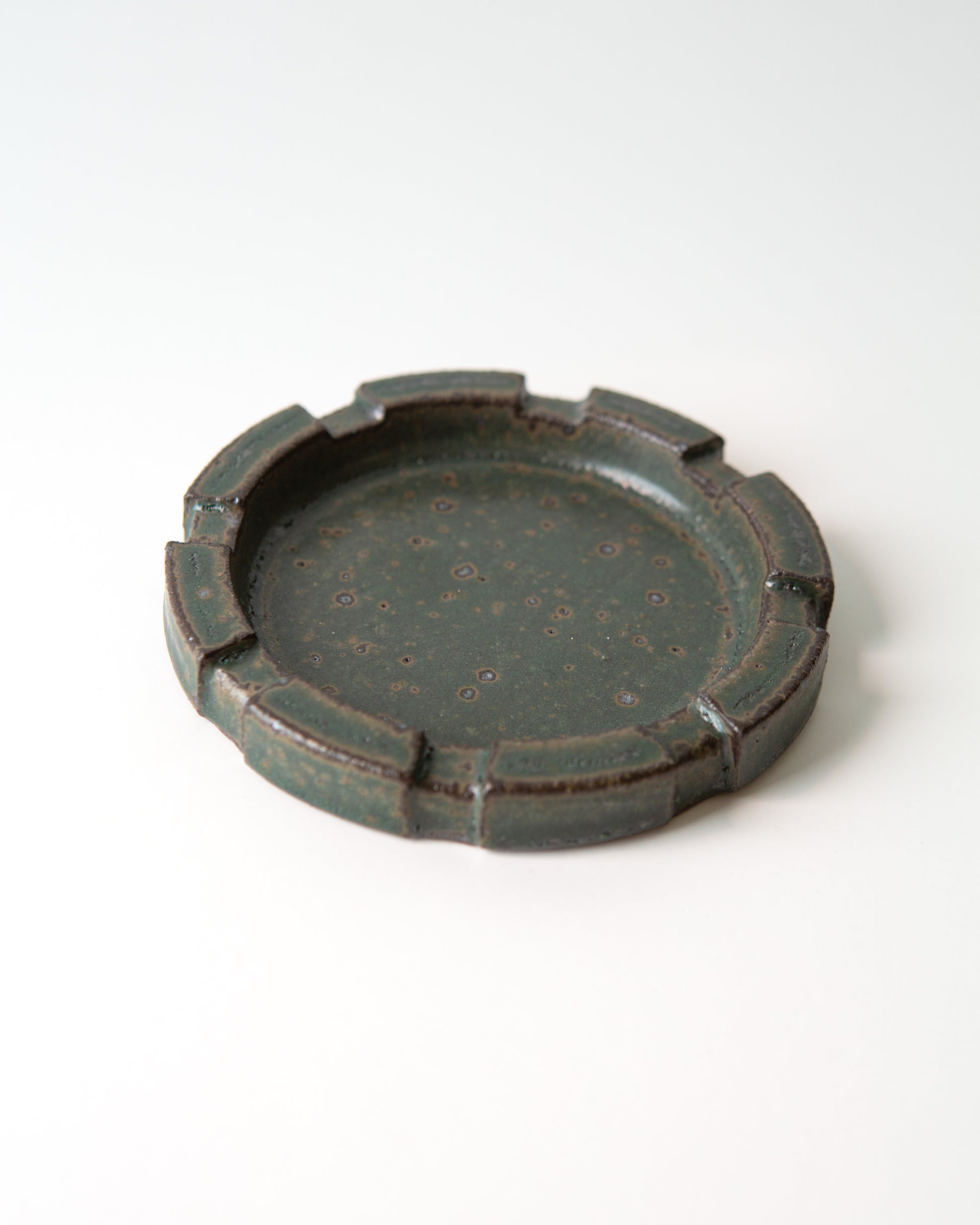 A round ceramic ashtray with a brutalist carved rim and a speckled dark green glaze, shown on a clean white background.