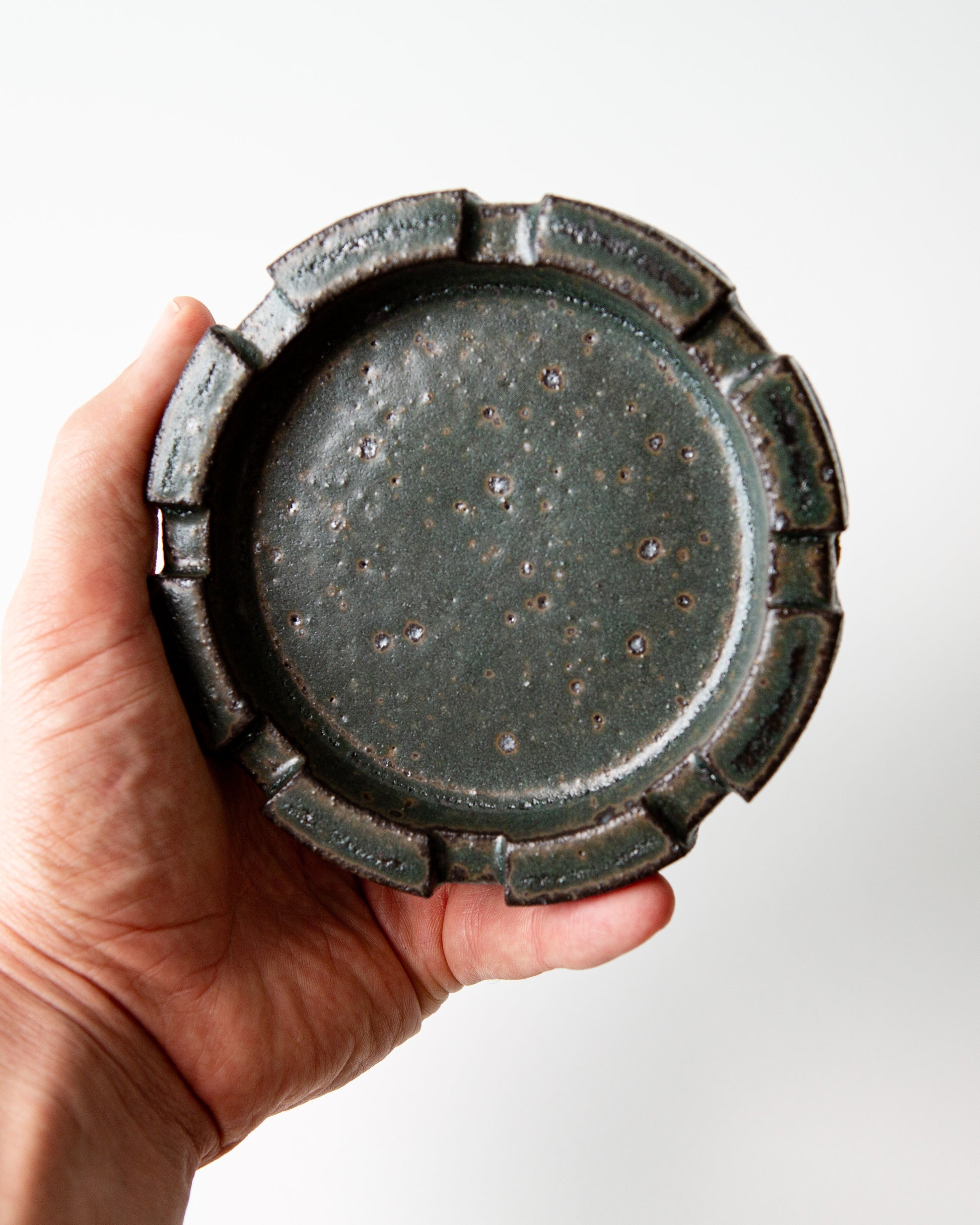 A hand holding a round ceramic ashtray with a brutalist carved rim and a speckled dark green glaze, shown on a clean white background.