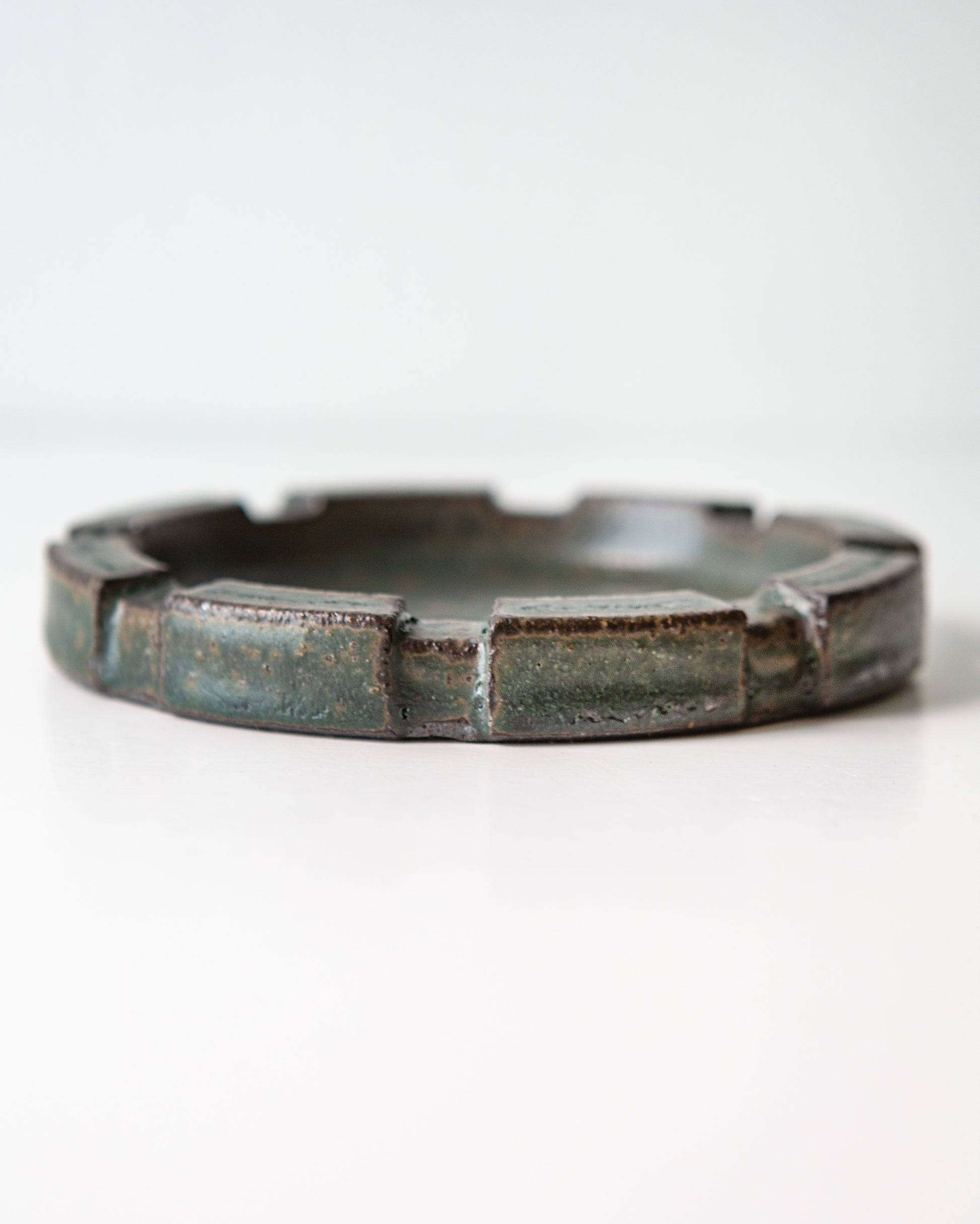 A close up shot of the side of a round ceramic ashtray with a brutalist carved rim and a speckled dark green glaze, shown on a clean white background.