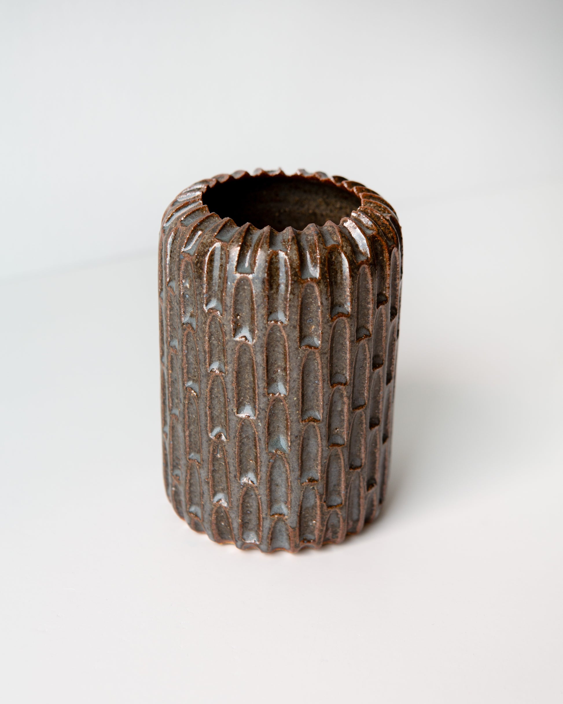 A small cylindrical ceramic vase with a textured, ribbed exterior and a flared rim, featuring a dark blue glaze with subtle coppery undertones, set against a plain white background.