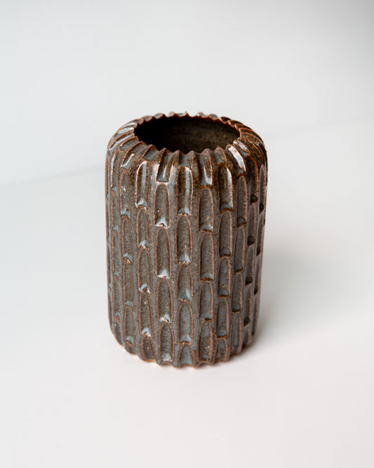 A small cylindrical ceramic vase with a textured, ribbed exterior and a flared rim, featuring a dark blue glaze with subtle coppery undertones, set against a plain white background.