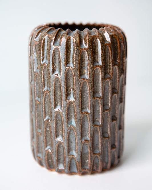 A close up on a small cylindrical ceramic vase with a notched exterior and a flared rim, featuring a dark blue glaze with subtle coppery undertones, set against a plain white background.
