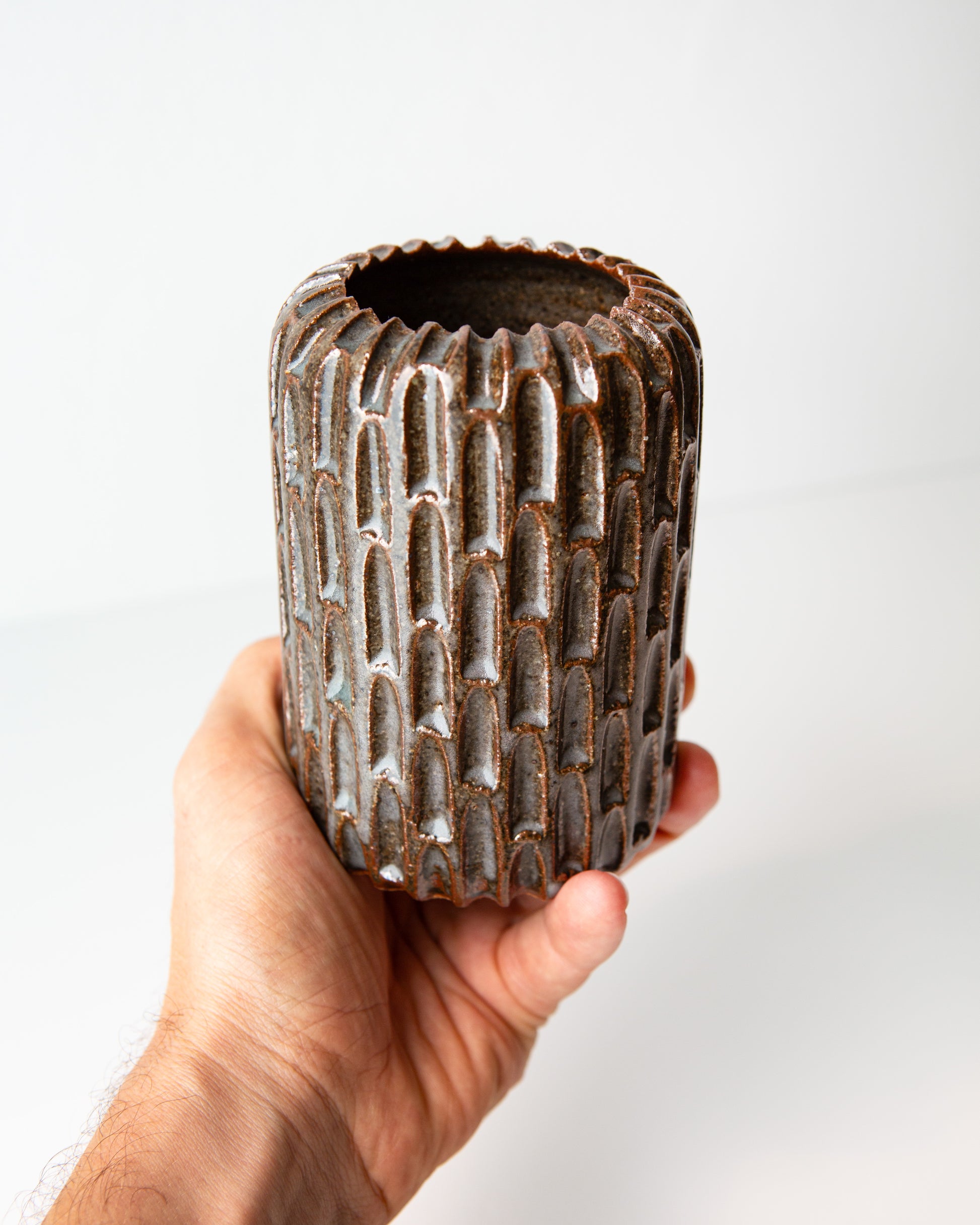 A hand holding a small cylindrical ceramic vase with a notched exterior and a flared rim, featuring a dark blue glaze with subtle coppery undertones, set against a plain white background.