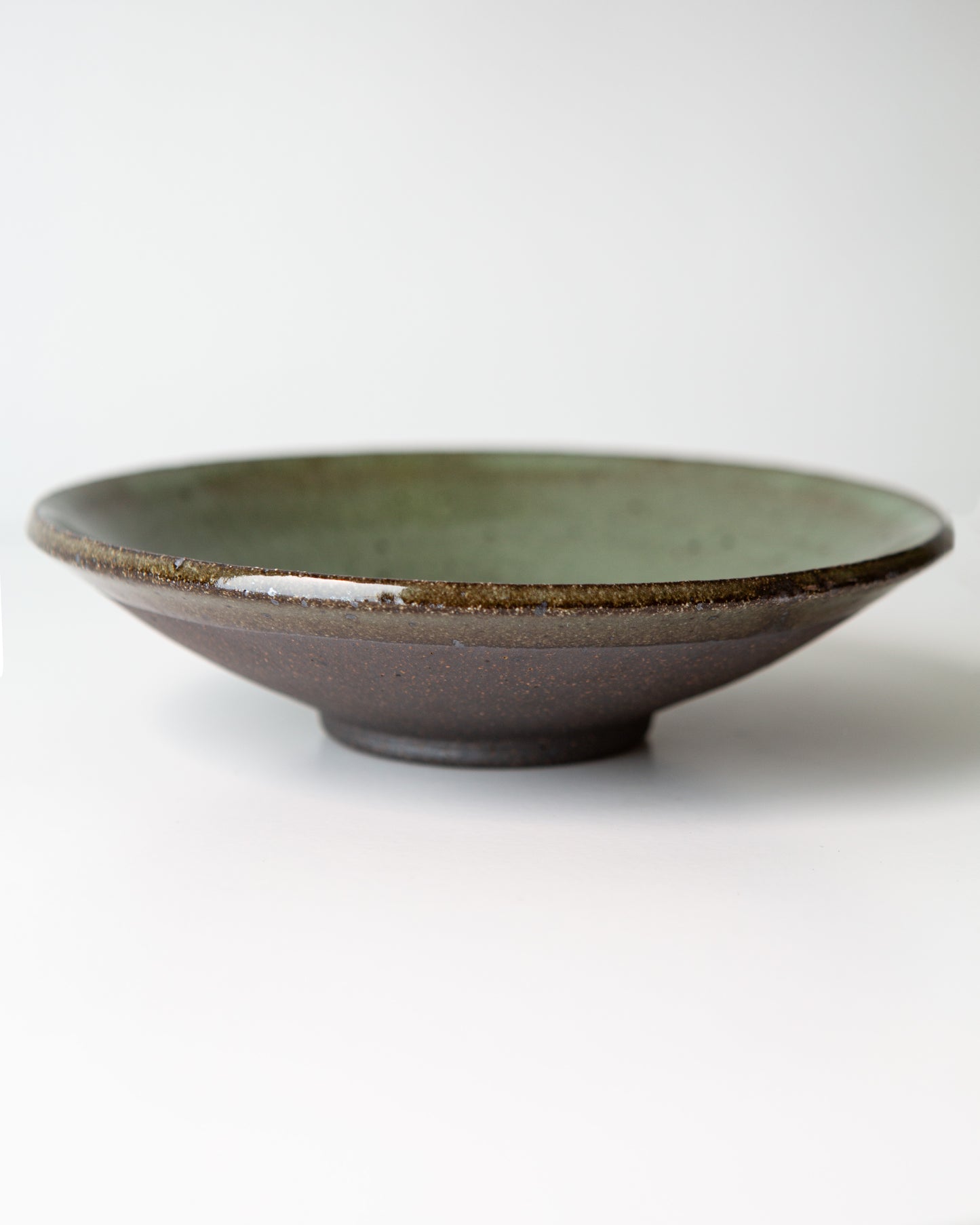 A handmade ceramic bowl with a dark brown exterior and a glossy olive green glazed interior, photographed on a clean white background.