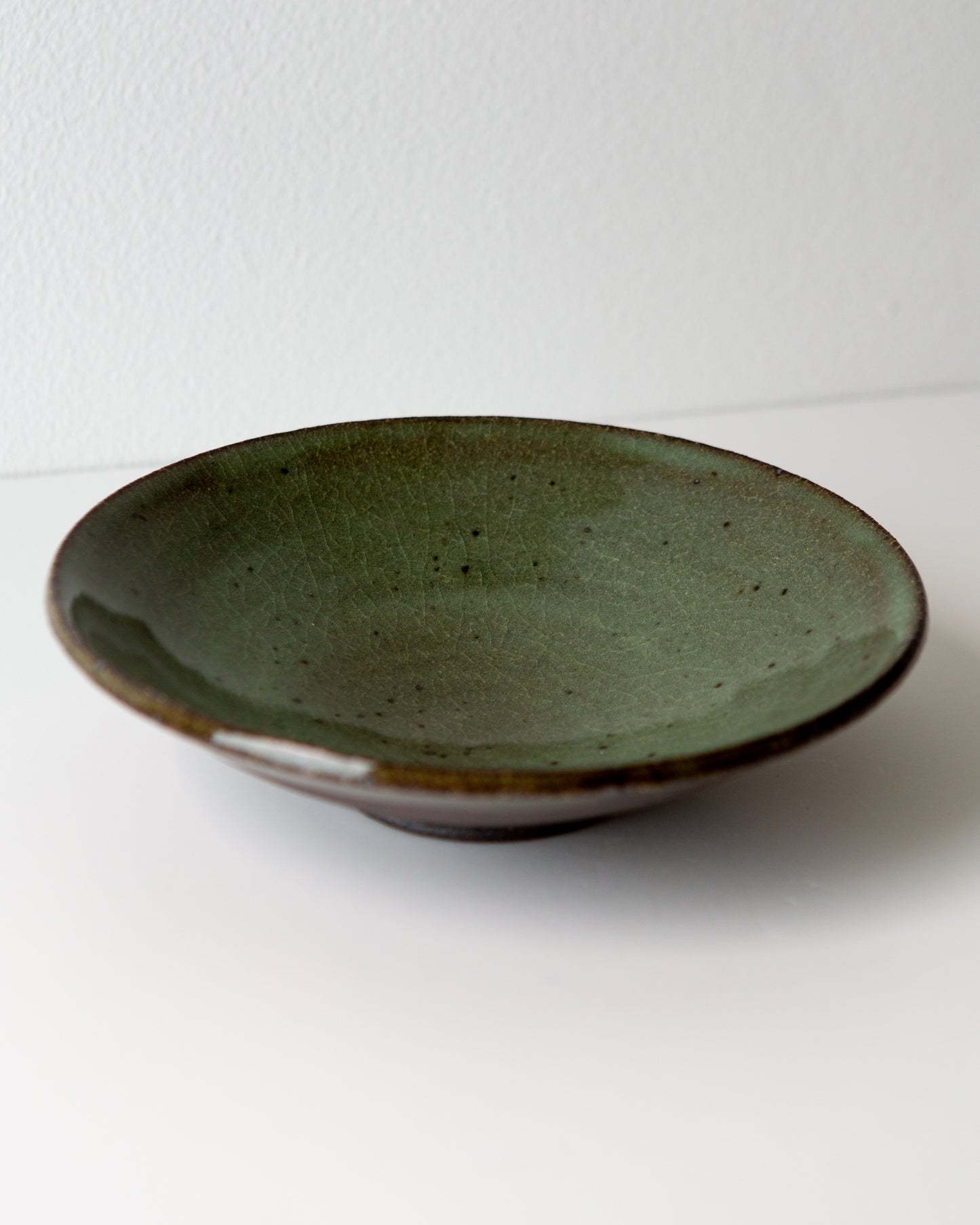 A handmade ceramic shallow bowl with a olive green glaze and a contrasting brown rim, photographed on a plain white surface against a white wall.