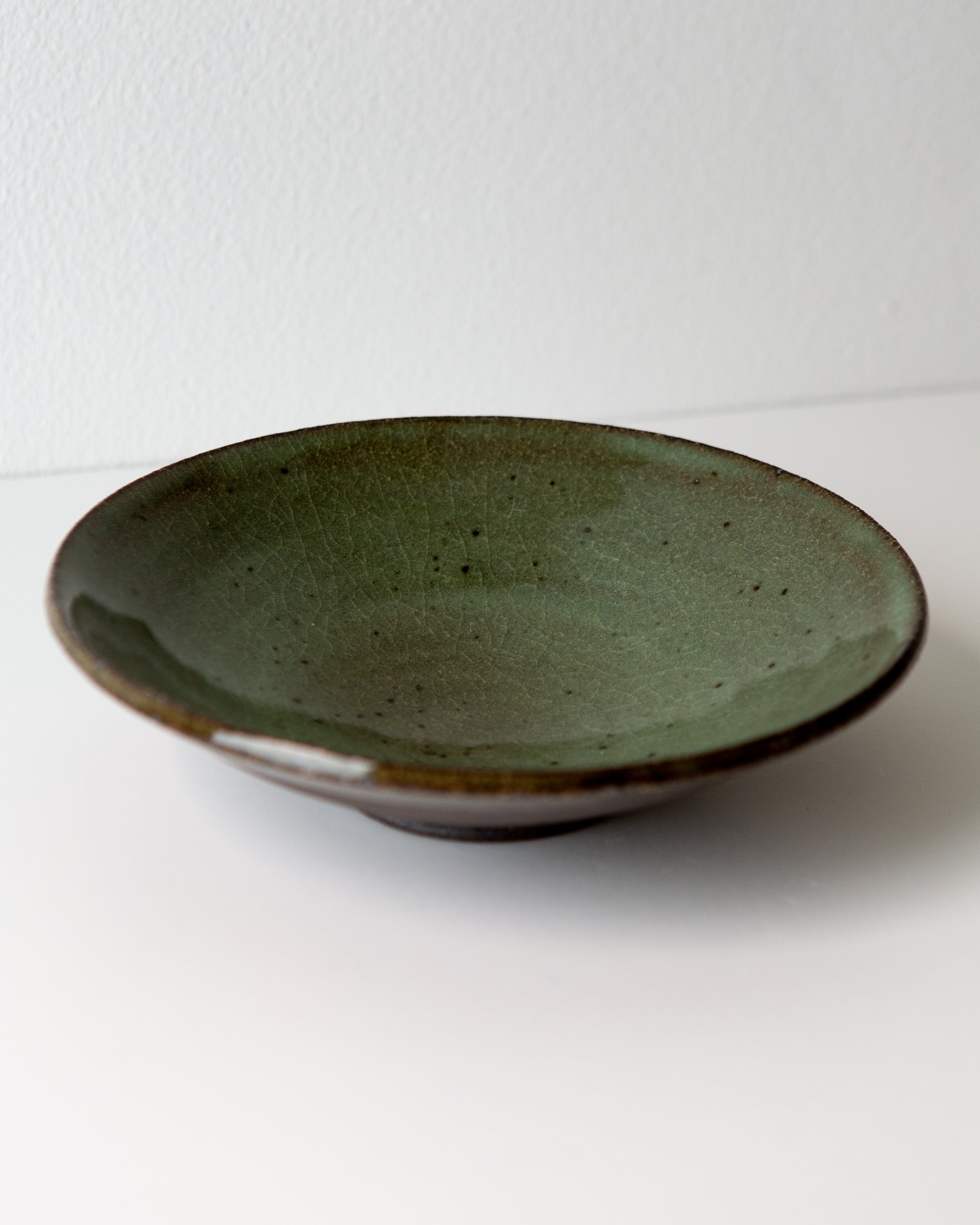 A handmade ceramic shallow bowl with a olive green glaze and a contrasting brown rim, photographed on a plain white surface against a white wall.