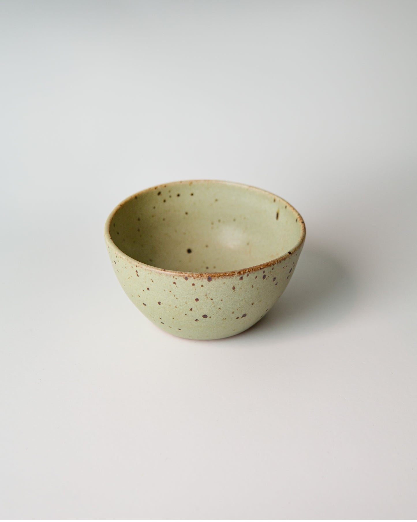 A small handmade ceramic bowl with a light teal green glaze and brown speckled details, photographed on a clean white background.