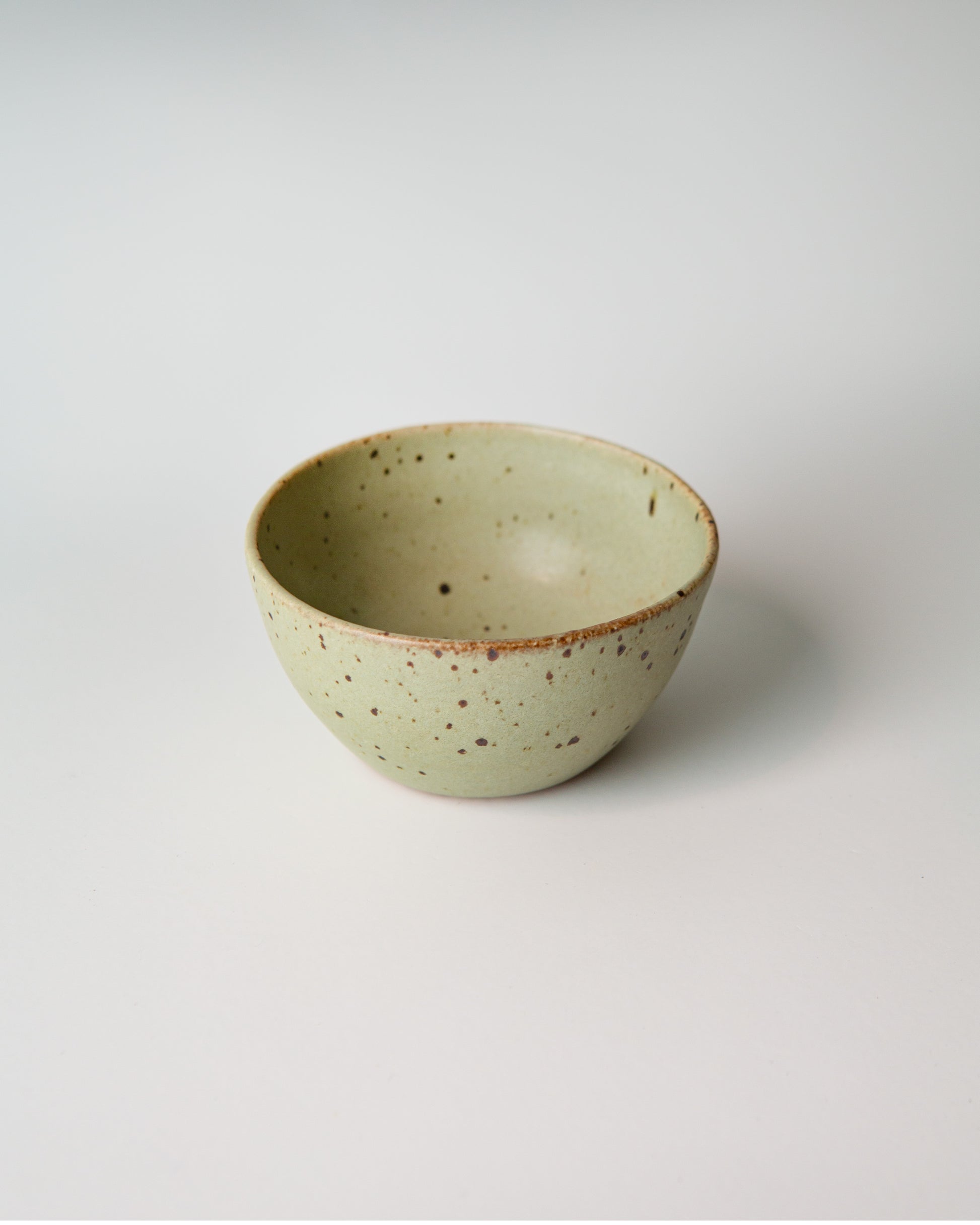 A small handmade ceramic bowl with a light teal green glaze and brown speckled details, photographed on a clean white background.