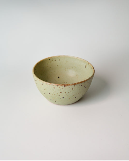 A small handmade ceramic bowl with a light teal green glaze and brown speckled details, photographed on a clean white background.