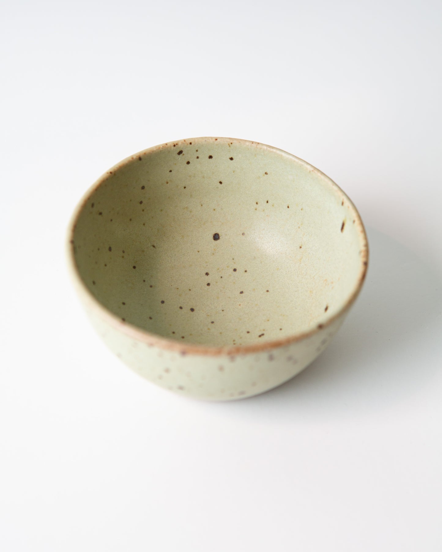 A small handmade ceramic bowl with a light teal green glaze and brown speckled details, photographed on a clean white background.