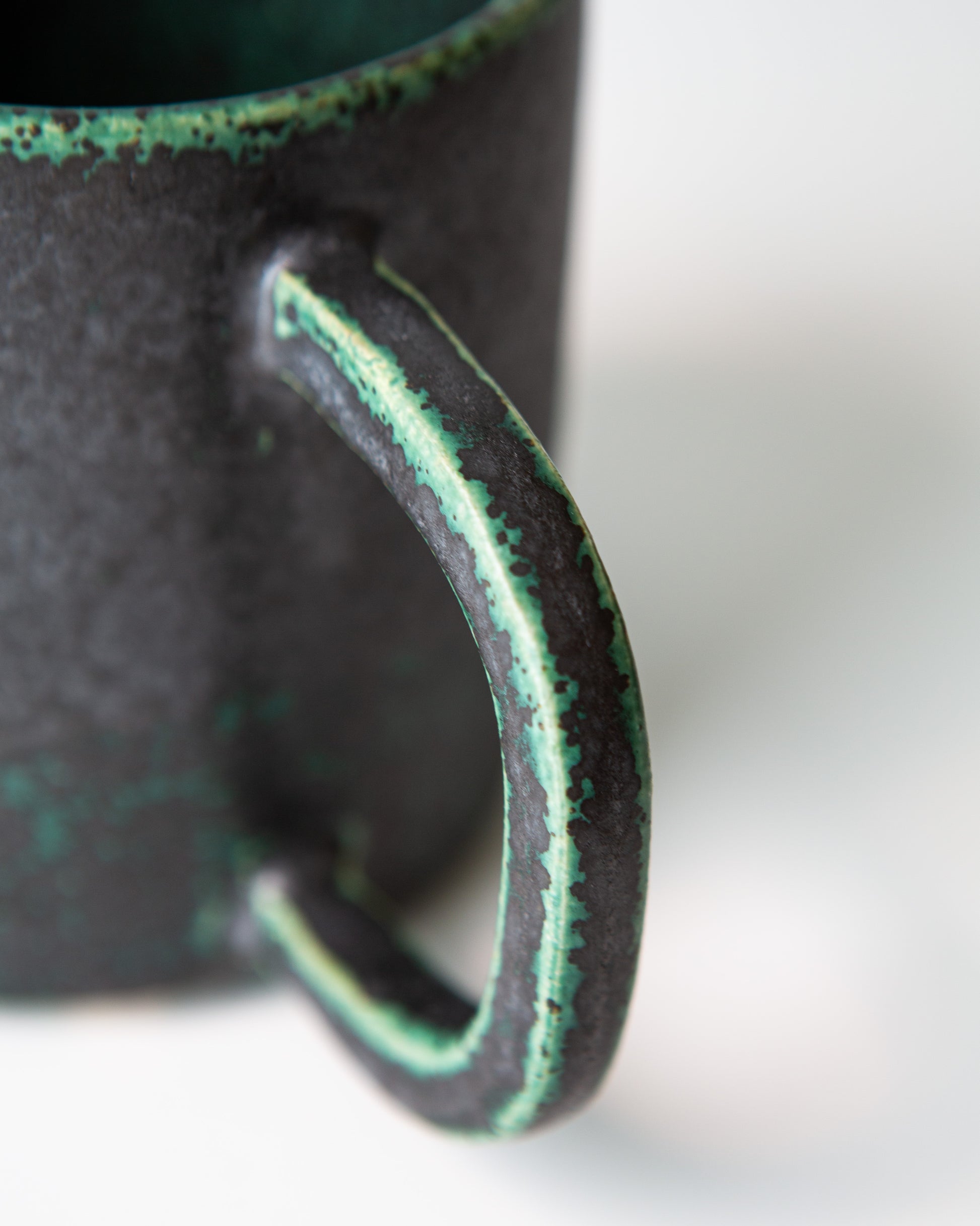 A close up shot of the handle of the ceramic mug with a textured matte black exterior, and a dark green glaze coloring on the edges, shown on a white background.