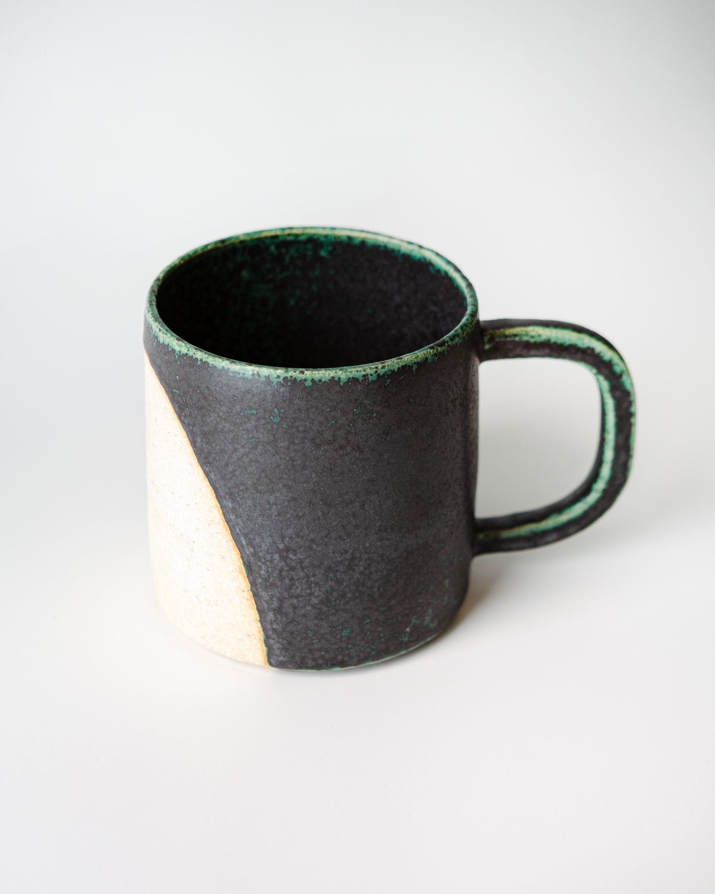 A handmade ceramic mug with a textured matte black exterior, a contrasting cream-colored unglazed half circle circle design shows on the lower left side, and a dark green glaze coloring on the edges, shown on a white background.
