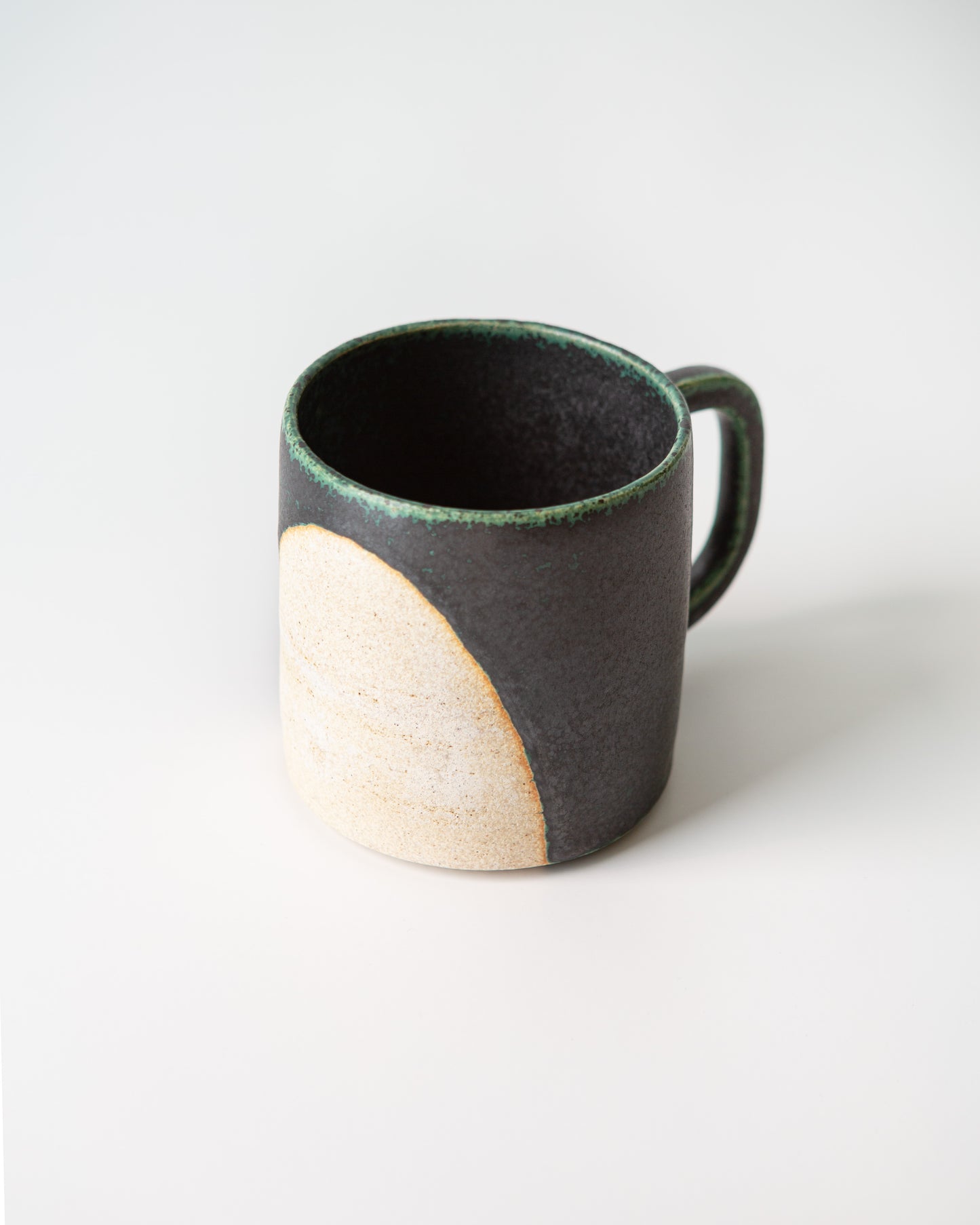 A second angle of the handmade ceramic mug with a textured matte black exterior, a contrasting cream-colored unglazed half circle circle design shows on the left side, and a dark green glaze coloring on the edges, shown on a white background.