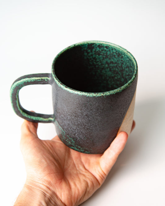 A handmade ceramic mug with a dark charcoal exterior and a speckled green glaze, held in a person's hand against a plain white background.