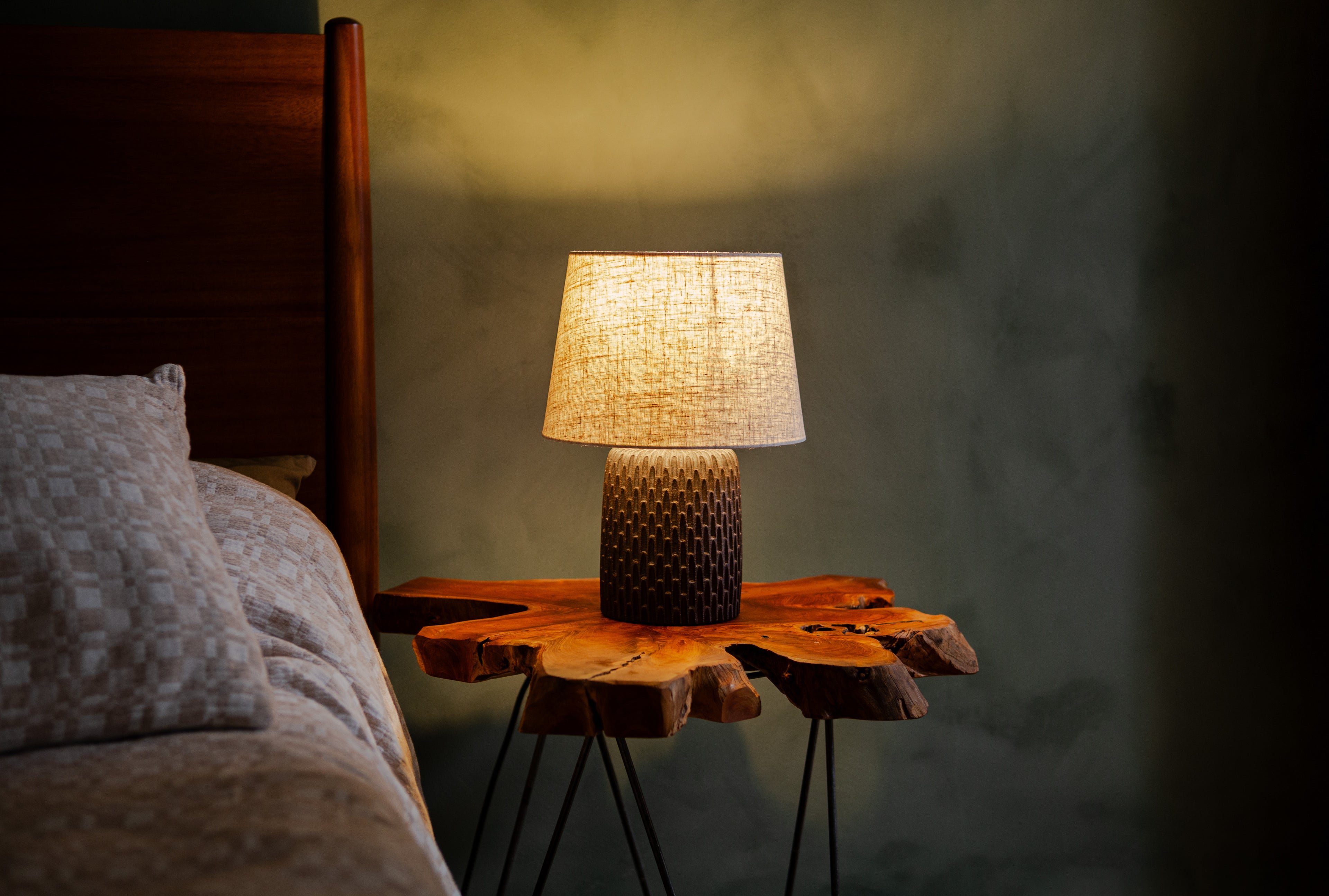 A table lamp with a hand carved dark brown ceramic base and a beige woven shade sits on a rustic live-edge wood nightstand beside a bed in a dimly lit bedroom.