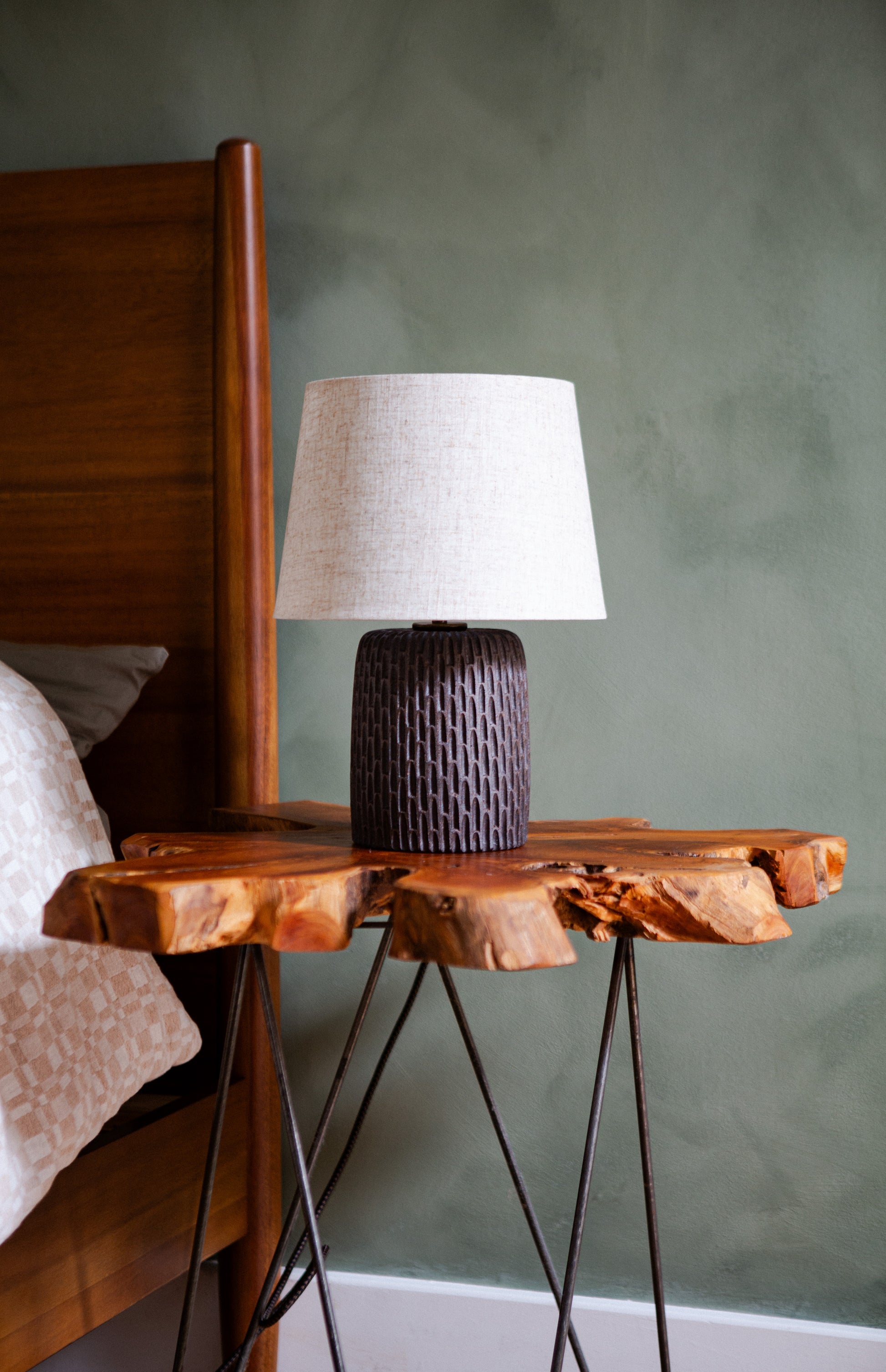 A dark brown hand carved table lamp with an off-white lamp shade, sitting on a wooden side table against a green wall.