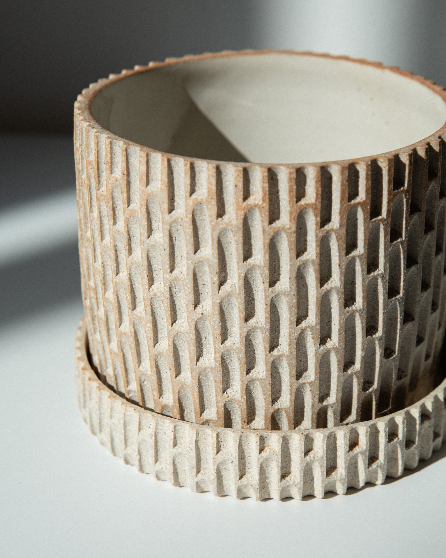 A close-up of a cylindrical ceramic planter with a notched exterior and a smooth, matte off-white interior, resting on a light surface with soft shadows.