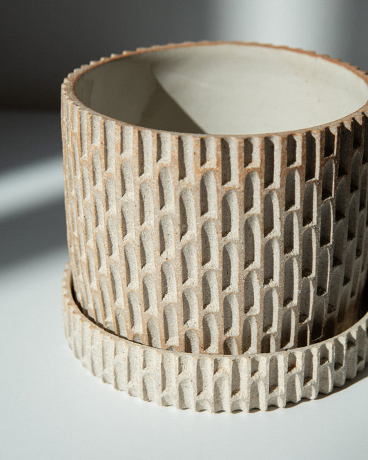A close-up of a cylindrical ceramic planter with a notched exterior and a smooth, matte off-white interior, resting on a light surface with soft shadows.