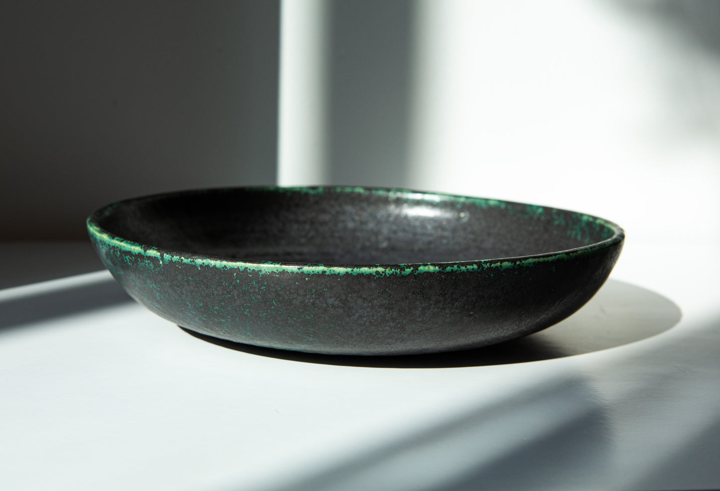A shallow round ceramic bowl with a dark charcoal-black glaze and a textured green rim, photographed in dramatic natural sunlight on a white surface.