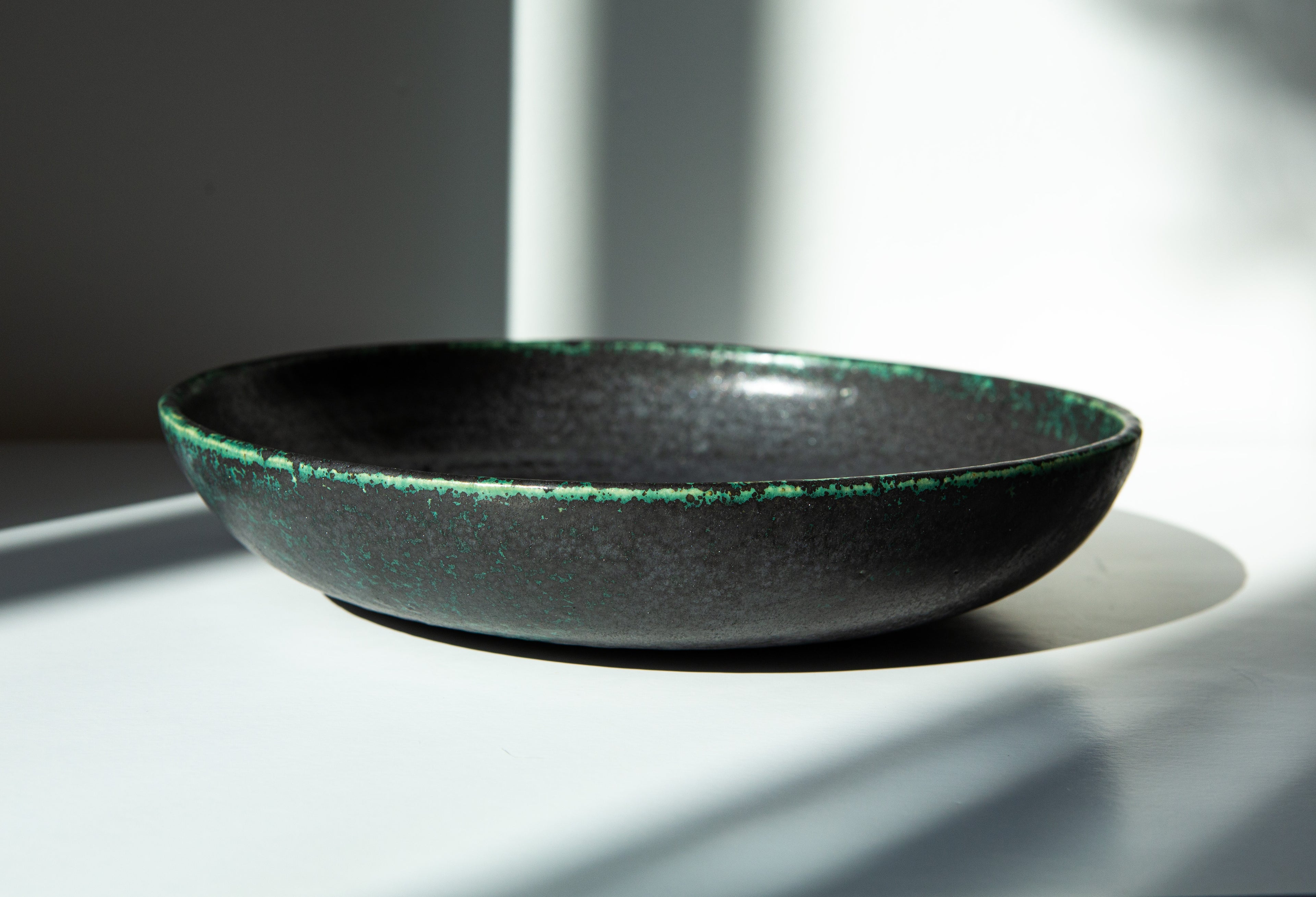 A shallow round ceramic bowl with a dark charcoal-black glaze and a textured green rim, photographed in dramatic natural sunlight on a white surface.