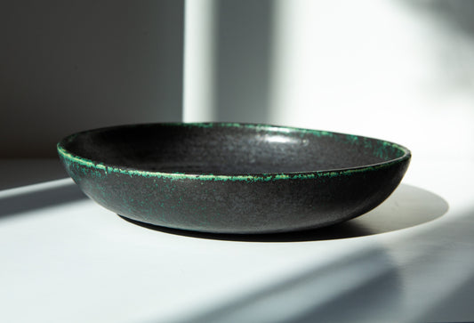 A shallow round ceramic bowl with a dark charcoal-black glaze and a textured green rim, photographed in dramatic natural sunlight on a white surface.