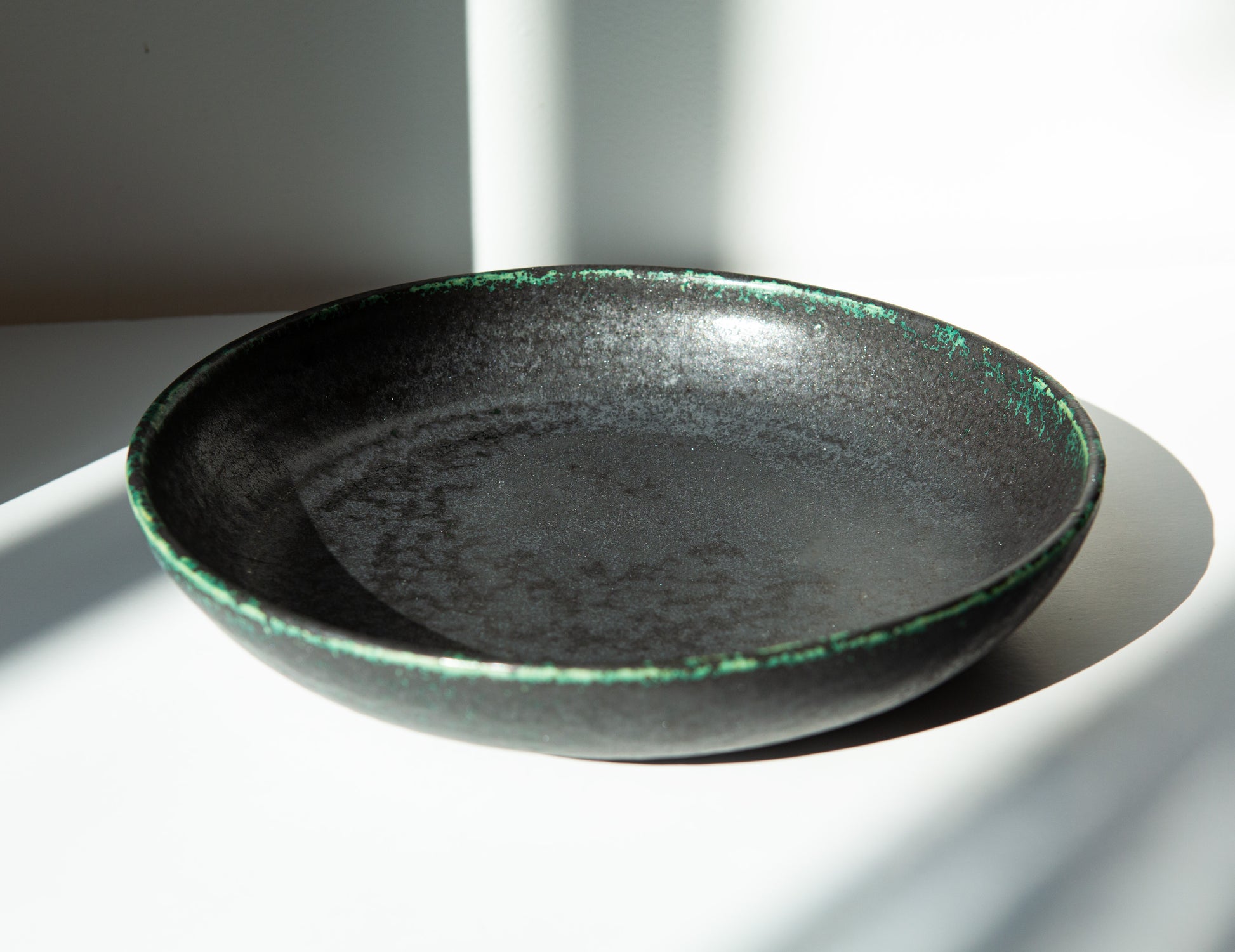 A shallow ceramic bowl with a black and green glaze, photographed in dramatic direct sunlight on a white surface.